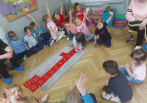 Zdjęcie przedstawia dzieci siedzące w kole, wokół szorstkich liczb, zgłaszające się do odpowiedzi poprzez podniesioną rękę.