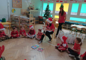 Zdjęcie przedstawia dzieci oraz panią dyrektor siedzące w kole. Pani dyrektor mówi do dzieci z wykorzystaniem maskotki renifera. Wszyscy ubrani są mikołajkowo.