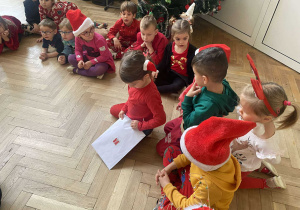 Zdjęcie przedstawia dzieci ubrane mikołajkowo. Dzieci siedzą na podłodze, jedno z nich otwiera kopertę.