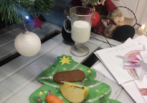 Zdjęcie przedstawia talerzyk w kształcie choinki, na którym leżą ciasteczko, kawałek jabłka oraz marchewki. Obok talerzyka stoi kubek z mlekiem.