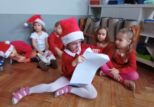 Zdjęcie przedstawia dzieci ubrane mikołajkowo. Dzieci siedzą na podłodze, jedno z nich otwiera kopertę.