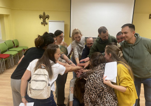 Zdjęcie przedstawia grupę osób stojących w kole. Rodzice mają wyciągniętą rękę przed siebie . Układają ręce jedna na drugą.