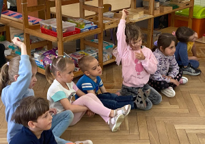 Zdjęcie przedstawia dzieci siedzące obok siebie. Dwoje dzieci zgłasza się do odpowiedzi.