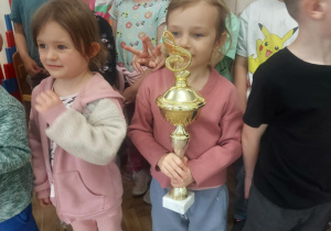 Na zdjęciu dziewczynka, która trzyma puchar, a w tle są dzieci