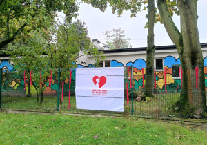 Zdjęcie przedstawia baner powieszony na ogrodzeniu przedszkola. Baner informuje o przystąpieniu przedszkola do kampanii Dzieciństwo bez przemocy.