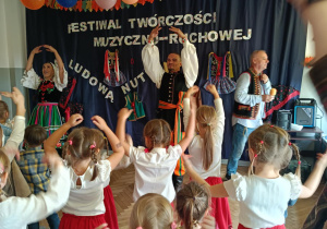 Zdjęcie przedstawia dzieci biorące udział w zabawie muzycznej prowadzonej przez osoby ubrane w stroje ludowe.