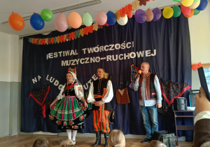 Zdjęcie przedstawia troje osób w strojach ludowych. Na drugim planie znajduje się dekoracja.