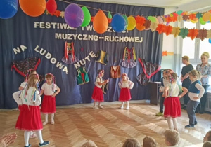 Zdjęcie przedstawia dzieci ubrane w stroje biało- czerwone. Dzieci prezentują taniec ludowy.