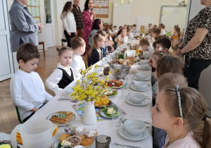 Zdjęcie przedstawia dzieci siedzące przy ogromnym wielkanocnym stole. Wokół stoją zgromadzeni pracownicy przedszkola.
