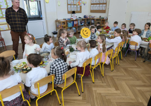 Zdjęcie przedstawia dzieci siedzące przy stole oraz pracownika przedszkola, który stoi obok.
