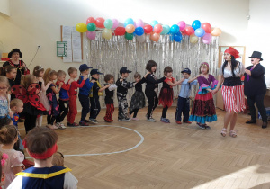 Zdjęcie przedstawia dzieci i nauczycielkę ustawionych w pociąg. W tle znajduje się dekoracja z balonów i srebrnej kurtyny. Tańcowi przyglądają się inne dzieci i nauczyciele.