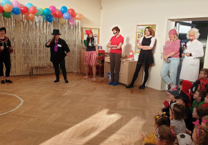 Zdjęcie przedstawia nauczycielki i dyrektora przebranych w stroje karnawałowe przemawiających do dzieci i innych uczestników balu. W tle znajduje się dekoracja z balonów i srebrnej kurtyny.