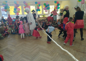 Zdjęcie przedstawia dzieci i nauczycieli przebranych w stroje karnawałowe. Biorą oni udział w zabawie tanecznej z wykorzystaniem liny.