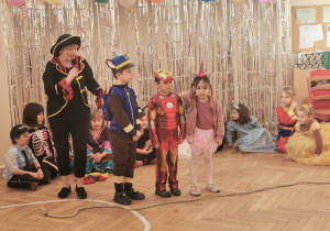 Zdjęcie przedstawia nauczycielkę prowadzącą imprezę oraz troje dzieci. Nauczycielka tłumaczy dzieciom przebieg zabawy. W tle znajduje się dekoracja z balonów i srebrnej kurtyny oraz siedzące dzieci.