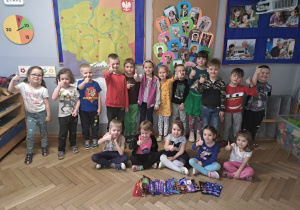 Zdjęcie przestawia grupę dzieci, które uśmiechają się do zdjęcia, wyciągając przed siebie dłonie z kciukami w górę. Przed dziećmi leżą czekolady.