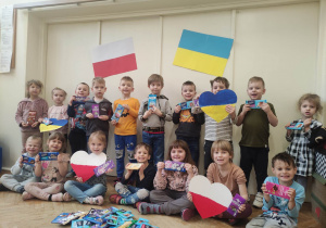 Zdjęcie przedstawia dzieci pozujące do fotografii, trzymające w rękach czekolady. Przed dziećmi na podłodze znajdują się również czekolady. W tle wiszą flagi: Polski i Ukrainy. Dzieci trzymają również dwa serca w barwach biało- czerwonych oraz dwa serca w barwach niebiesko- żółtych.