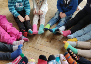 Zdjęcie przedstawia dzieci siedzące w kole. Dzieci siedzą z wyciągniętymi przed siebie stopami, na których mają różnokolorowe skarpetki.