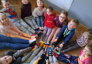 Zdjęcie przedstawia dzieci siedzące w kole. Dzieci mają wyciągnięte przed siebie stopy, na które założyli różnokolorowe skarpetki.