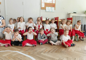 Zdjęcie przedstawia dzieci ubrane w białe bluzki i czerwone spodnie/ spódniczki. Wszyscy siedzą i pozują do zdjęcia.