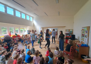 Zdjęcie przedstawia nauczycieli tańczących w kole. Tańcowi przyglądają się dzieci siedzące na podłodze.