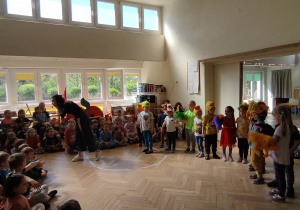 Zdjęcie przedstawia dzieci ustawione w rzędzie. Dzieci mają maski różnych ptaków. Ich zabawie przyglądają się dzieci siedzące na podłodze. Przed dziećmi stoi prowadząca koncert śpiewająca do nich.