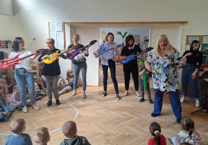 Zdjęcie przedstawia nauczycieli grających na balonowych gitarach oraz osobę prowadzącą koncert.