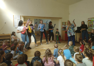 Zdjęcie przedstawia nauczycieli grających na balonowych gitarach oraz osobę prowadzącą koncert. Zabawie przyglądają się dzieci siedzące na podłodze.