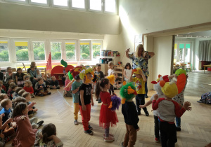 Zdjęcie przedstawia dzieci w maskach ptaków biorące udział w zabawie muzyczno- ruchowej z prowadzącą. Na podłodze siedzą dzieci i przyglądają się zabawie.