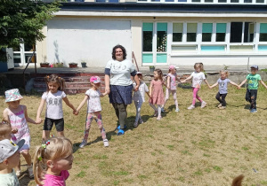 Zdjęcie przedstawia dzieci i nauczycielkę tańczących w kole w ogrodzie.