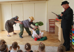 Zdjęcie przedstawia pracowników przedszkola w strojach, którzy odgrywają utwór "Rzepka". Przedstawienie oglądają zgromadzone w sali dzieci.