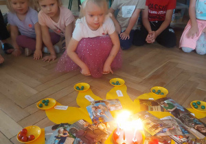 Zdjęcie przedstawia dziewczynkę, która dmucha świeczki na torcie urodzinowym, w tle widać siedzące dzieci