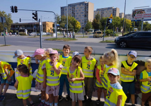 Na zdjęciu widzimy grupkę dzieci, które stoją na skrzyżowaniu i obserwują poruszające się pojazdy.