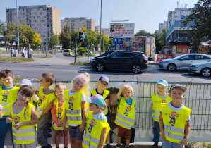 Na zdjęciu widzimy grupkę dzieci, które stoją na skrzyżowaniu i obserwują poruszające się pojazdy.