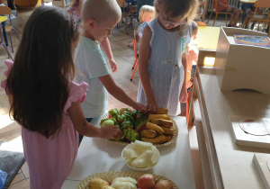 Na zdjęciu widzimy trójkę dzieci, która częstuje się owocami przyniesionymi przez solenizantkę na urodziny.