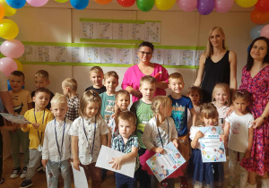 Zdjęcie przedstawia grupę dzieci wraz z dyrektorem przedszkola i nauczycielkami grupy. Wszyscy pozują do zdjęcia na tle dekoracji z napisem dzień przedszkolaka oraz kolorowymi balonami.
