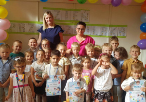 Zdjęcie przedstawia grupę dzieci wraz z dyrektorem przedszkola i nauczycielką grupy. Wszyscy pozują do zdjęcia na tle dekoracji z napisem dzień przedszkolaka oraz kolorowymi balonami.