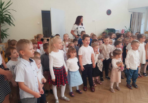 Zdjęcie przedstawia dzieci z całego przedszkola i nauczyciela śpiewających hymn przedszkolaka.
