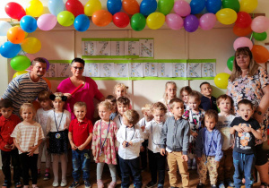 Zdjęcie przedstawia grupę dzieci wraz z dyrektorem przedszkola i nauczycielami grupy. Wszyscy pozują do zdjęcia na tle dekoracji z napisem dzień przedszkolaka oraz kolorowymi balonami.