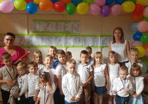 Zdjęcie przedstawia grupę dzieci wraz z dyrektorem przedszkola i nauczycielką grupy. Wszyscy pozują do zdjęcia na tle dekoracji z napisem dzień przedszkolaka oraz kolorowymi balonami.