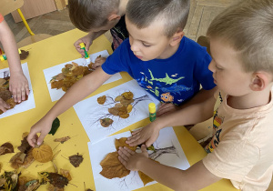 Dzieci siedzą przy stole i przykleją liście do konturu drzewa.