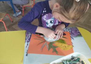 Na zdjęciu widzimy dziewczynkę, która rysuje kredkami pastelowymi nos jeżykowi stworzonemu z liści.