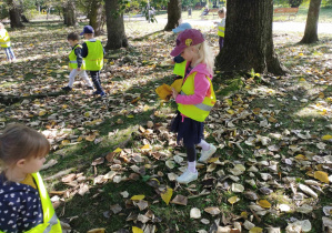 Na zdjęciu widzimy grupkę dzieci, które zbierają liście z ziemi w pobliskim parku.