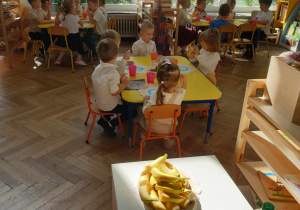 Na zdjęciu widzimy grupkę dzieci, które siedzą przy stolikach i jedzą słodkości.