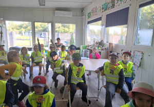 Zdjęcie przedstawia dzieci siedzące w sali. Dwoje z nich podnosi rękę, by zgłosić się do odpowiedzi.
