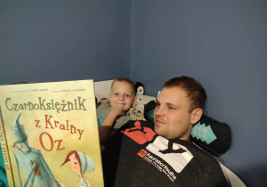 Zdjęcie przedstawia mężczyznę, czytającego chłopcu książkę.