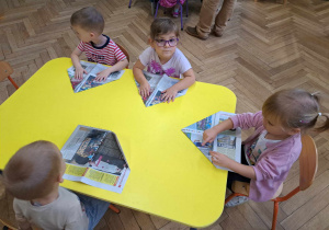 Zdjęcie przedstawia dzieci siedzące przy stole i składające gazetę.