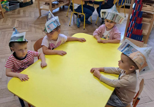 Zdjęcie przedstawia dzieci siedzące przy stole, w czapkach papierowych na głowie.