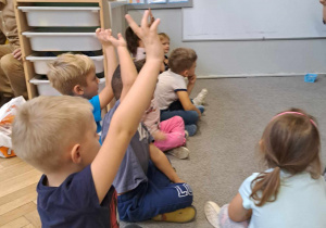 Zdjęcie przedstawia dzieci siedzące na dywanie i zgłaszające się do odpowiedzi.