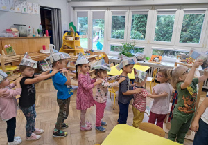 Zdjęcie przedstawia dzieci stojące jedno za drugim, w papierowych czapkach na głowie.