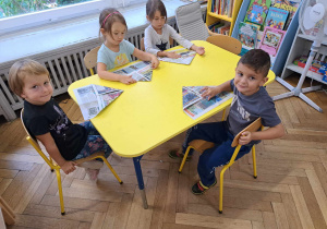 Zdjęcie przedstawia dzieci siedzące przy stole i składające gazetę.
